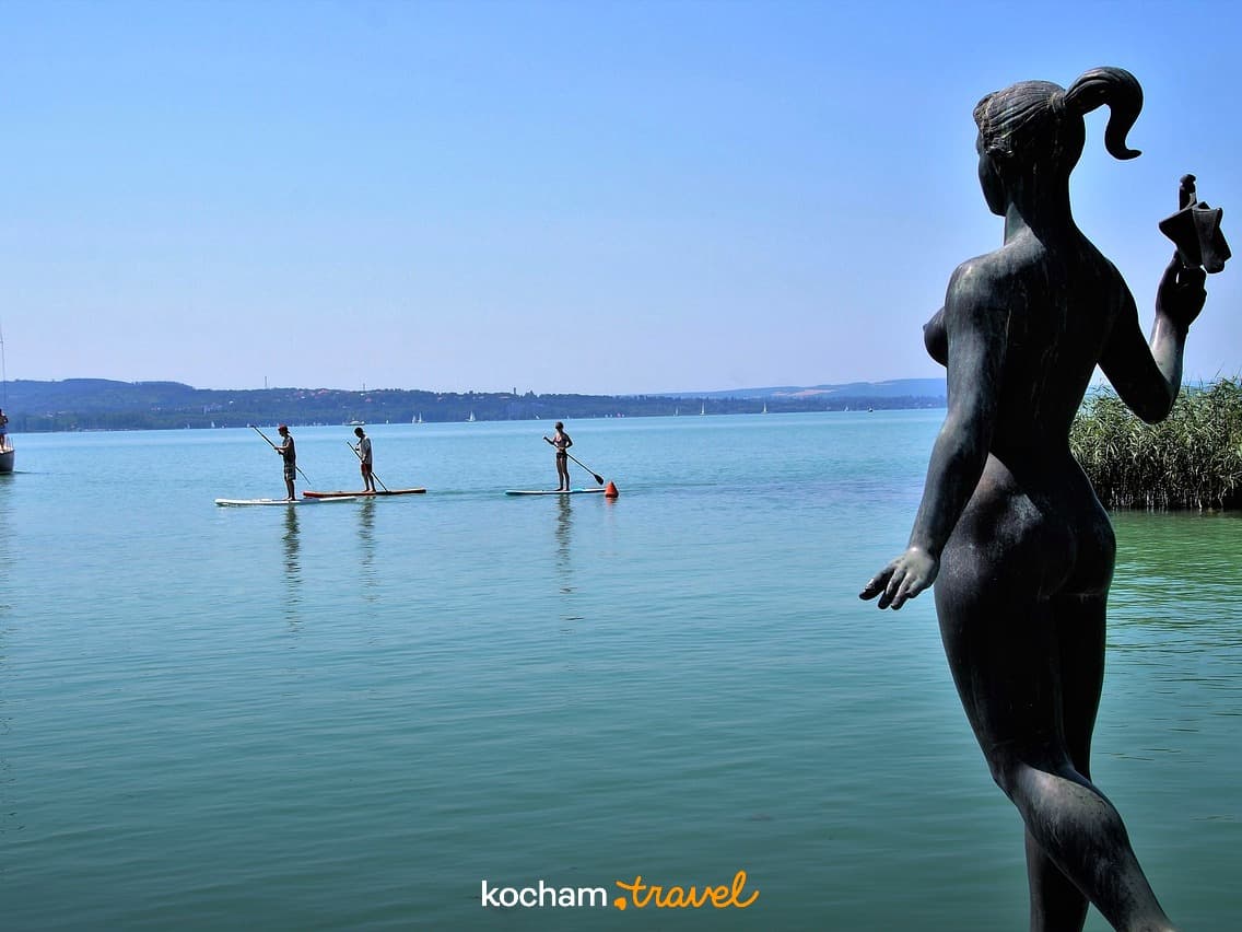 Posąg kobiety, paddleboardziści, żaglówka na turkusowym jeziorze.