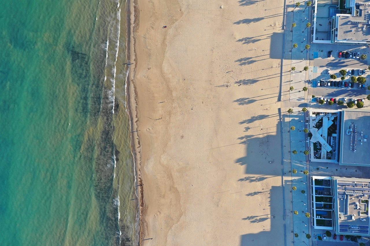 Widok z lotu ptaka: piaszczysta plaża, turkusowa woda, palmy, budynki, cienie.