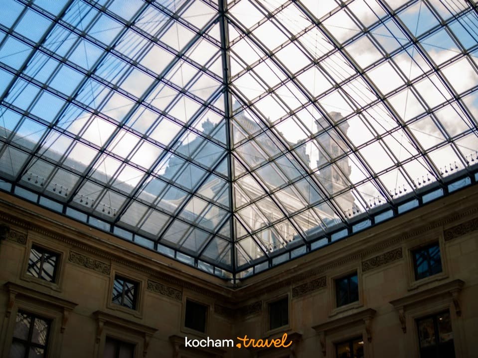 Szklany świetlik w kształcie piramidy, odbijający klasyczną architekturę i niebo.
