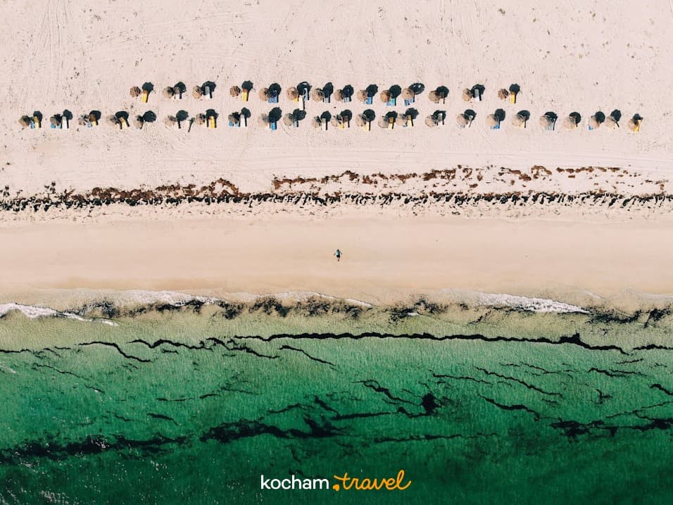 Widok z góry: tropikalna plaża, parasole, turkusowa woda, spacerująca osoba.