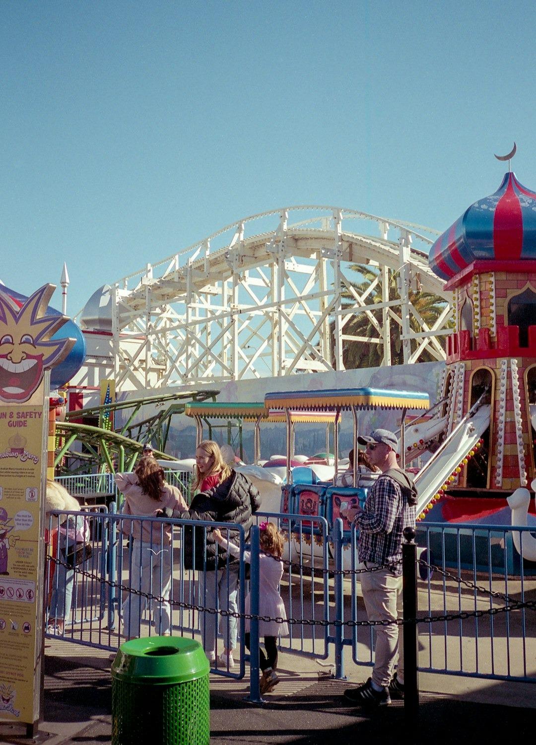 Ludzie w wesołym miasteczku, roller coaster, kolorowe atrakcje, słoneczny dzień.