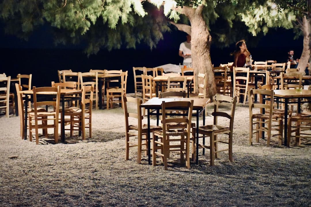 Zewnętrzna restauracja wieczorem, drewniane stoły, krzesła, drzewa, żwir.
