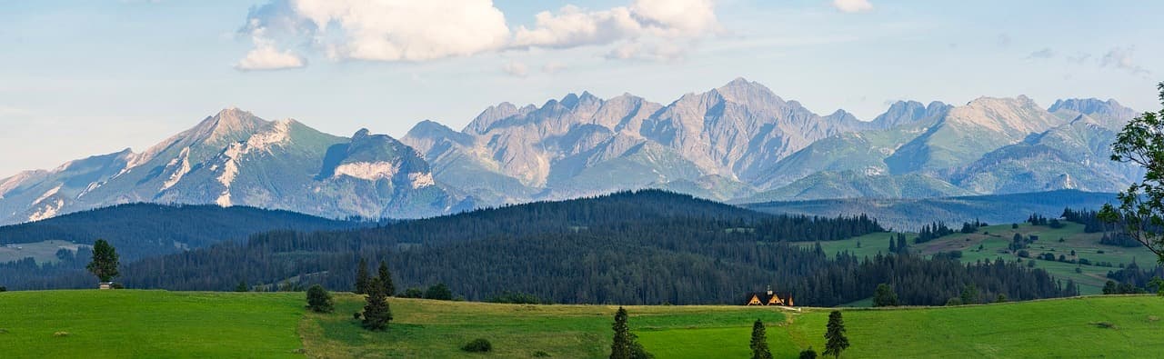 Panorama Tatr, zielone łąki, las, drewniany dom, błękitne niebo.