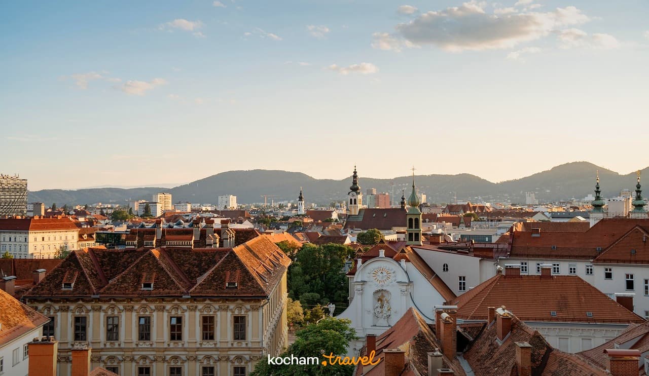 Panoramiczny widok: dachy europejskiego miasta, wieże kościelne, góry, złote światło.